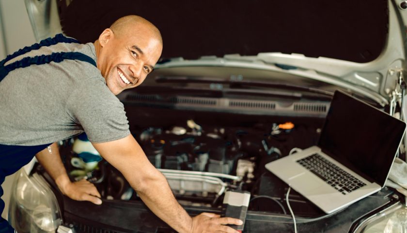 un homme souriant analysant le moteur de sa voiture avec un ordinateur portable